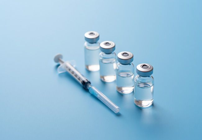 Четыре ампулы стоят в ряд на светло-голубом фоне рядом с медицинским шприцем